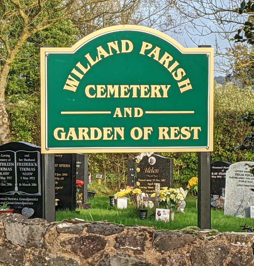 Sign depicting the entrance to the village cemetery on a green background with gold writing and a well kept graveyard in the background.